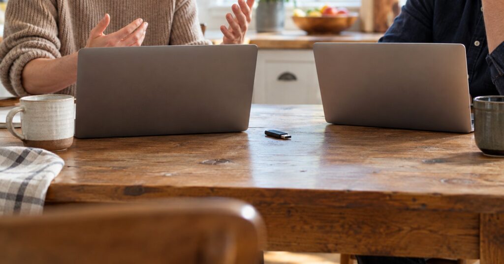 Two people talking at a kitchen table, with laptops turned away and a USB stick between them.